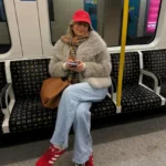 Woman sitting on subway train with smartphone, wearing a red cap and cozy coat.